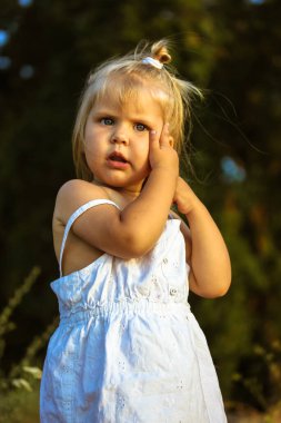 Şaşırmış ciddi çocuk. Yaz günü beyaz elbiseli, sarı saçlı, sevimli, sevimli, küçük, beyaz Avrupalı bir kız kameraya ilgiyle bakıyor. 2-4 yaşlarında bir çocuk, parkta yeni yürümeye başlamış..