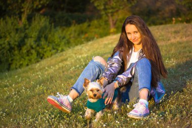 Kiev. Ukrayna. 1 Mayıs 2021. Gümüş renkli yağmurluk giymiş uzun saçlı genç bir kız, kot pantolon park çimlerinde, çayırda Yorkshire teriyeri, küçük bir köpek, köpek yavrusu. İnsanlar ve evcil hayvanlar, hayvan severler konsepti. 