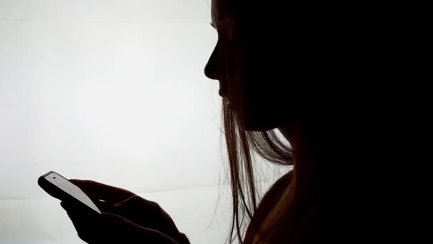 一个拿着智能手机在白色背景上滚动的女孩的轮廓 年轻女性形象地使用手机 在网上搜寻信息 发短信 网上购物 — 图库视频影像