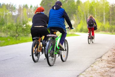 Kiev, Ukraine. April 7, 2021. Group of cyclists ride on an asphalt road in green forest, park at spring day. View from back of three people on bikes. Sporty way of life. Travel, trip, journey concept.