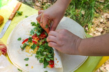 Burrito 'ya sarılı kolların üst görüntüsü, sebzeli pide ekmeği, otlar, peynir. İlkbaharda, yaz gününde doğada piknik. Parkta, ormanda yemek pişirmek. Yeşil sağlıklı vejetaryen yemeği. Hazırlanıyor.