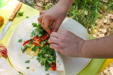Burrito 'ya sarılı kolların üst görüntüsü, sebzeli pide ekmeği, otlar, peynir. İlkbaharda, yaz gününde doğada piknik. Parkta, ormanda yemek pişirmek. Yeşil sağlıklı vejetaryen yemeği. Hazırlanıyor.