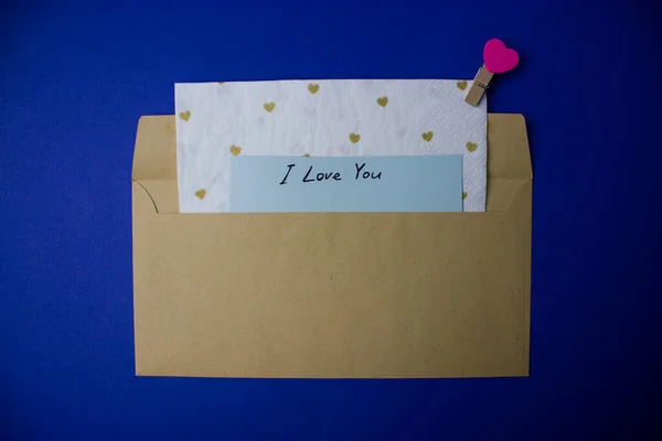 Brown envelope with blue note with text I love you on blue background top view. Congratulations for Valentine\'s Day. Confession of love, feelings concept. Decor in the shape of a pink heart, hearts