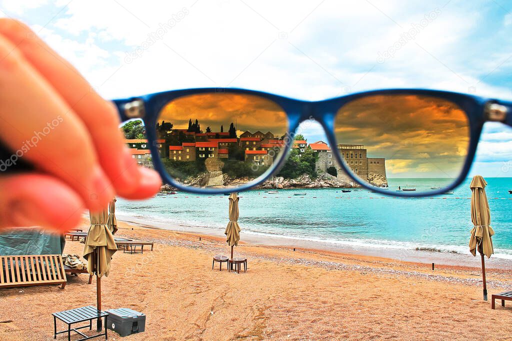 June 1, 2017. Budva, Montenegro. Sveti Stefan resort village, island. View of the ancient architecture through glasses of blue sunglasses. Deserted sandy beach with umbrellas, sun loungers. Summer sea