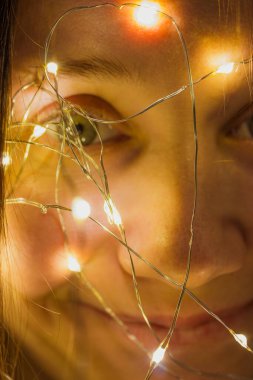 Gülümseyen kız parıldayan ışıklarla çelenklerin arasından bakıyor. Genç kadın 2022 yılbaşı arifesi için bir Noel ağacı süslemeye hazırlanıyor ve eğleniyor. Odaklanan ince metal tel. Dikey portre.