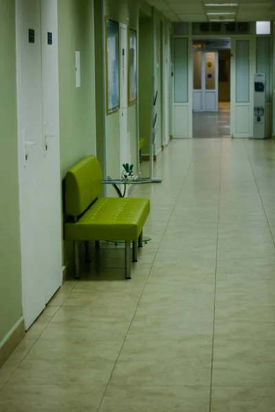 Kyjev, Ukrajina. 1. června2020. Zelená pohovka pro čekající pacienty v dlouhé prázdné nemocniční chodbě. Chodba v moderním zdravotnickém zařízení s bílými zavřenými dveřmi. Umělé osvětlení uvnitř. Interiér — Stock fotografie