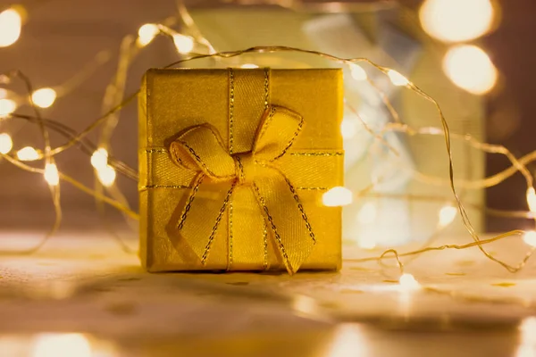 Scatola regalo gialla con cavalletti a prua sul tavolo tra luci a ghirlanda intrecciate luminose. Capodanno 2022, Natale. Magiche vacanze invernali shopping. Scatola d'oro con regalo la sera. Sfondo festivo — Foto Stock