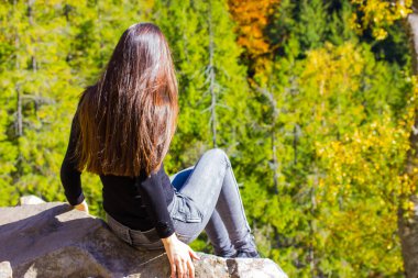 Dağın tepesinde uzun siyah saçlı bir kızın yeşil kozalaklı bir ormana karşı oturması. Kimliği belirsiz genç bir kadın ormanda tek başına yürüyüş yapıyor. Macera, gezi konsepti.