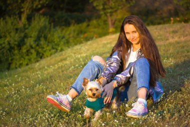 Kyiv. Ukrayna. 1 Mayıs 2021. Gümüş renkli yağmurluk giyen, kot pantolonlu, uzun saçlı genç bir kadın park çimlerinde Yorkshire teriyeri olan bir çayır, küçük bir köpek, köpek yavrusu. İnsanlar ve hayvanlar, hayvan severler dışarıda eğlenirler.