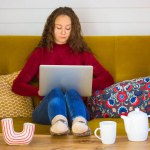 Pretty brunette girl is sitting on couch with laptop among colorful soft decorative pillows. Young woman working remotely, studying, shopping online at autumn winter. Wooden table with tea cup, teapot Pretty brunette girl is sitting on couch with laptop among colorful soft decorative pillows. Young woman working remotely, studying, shopping online at autumn winter. Wooden table with tea cup, teapot
