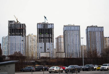Kyiv, Ukrayna. 8 Ocak 2022. Tamamlanmamış çok katlı apartmanlar, gri gökyüzüne karşı inşaat vinçleri. Yüksek binaların inşaatı. Şehir manzarası. Otoparka park etmiş arabalar.