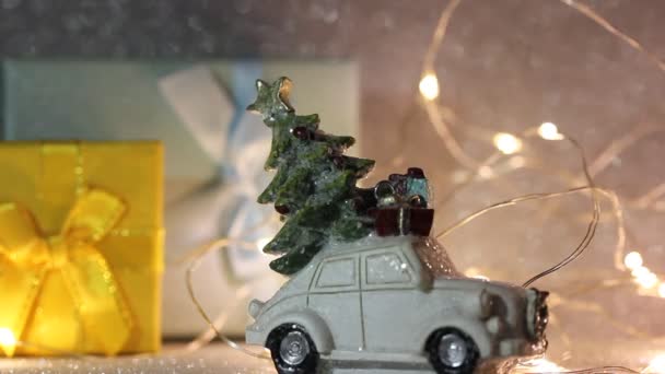 Brinquedo carro retro branco com árvore de Natal, caixas de presente no telhado. Ano Novo 2022, conceito de Natal. Compra de presentes, compras de inverno. Presentes, guirlanda luzes amarelas no fundo festivo brilhante prateado. — Vídeo de Stock