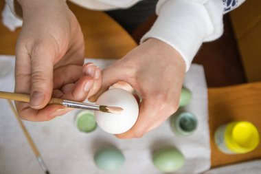 Kadının elinde boya fırçası ve temiz beyaz tavuk yumurtası. Kadın bej renkli üst manzara ile yumurta boyamaya hazırlanıyor. Paskalya hazırlığı konsepti. İnsanlar ve gelenekler. Arka planda yeşil yumurtalar
