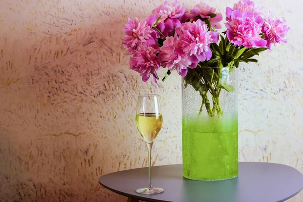緑の花瓶のピンク色の牡丹の花束は モダンなインテリアのテーブルの上に立って シャンパンのガラスが近くに立っています 家庭の温暖化 家庭の概念での休日 夏の花を咲かせます テキストの場所 — ストック写真