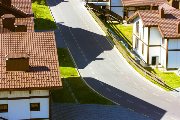 Aero view of brown tiled roofs with brick pipes of one-story, two-story houses paved street view from above. Green lawns with fresh grass and a asphalt road with markings goes into a bend at sunny day