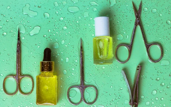 Manicure and pedicure items set on green background with water drops. Manicure scissors, tweezers, cutters, cuticle oil, nail growth oil. Beauty shop goods. Nail care cosmetic tools. Spa treatments.