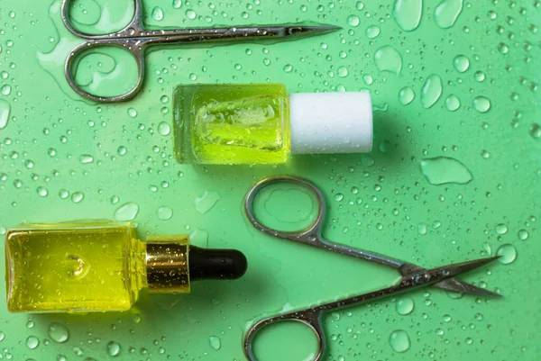 Manicure and pedicure items set on green background with water drops. Manicure scissors, cuticle oil in bottles, nail growth oil. Beauty shop goods. Nail care cosmetic tools. Spa treatments concept.