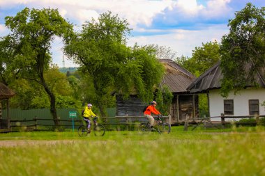 Kyiv, Ukrayna. 1 Mayıs 2021 İki bisikletçi yeşil bir bahar çayırında bisiklet sürüyor. Arka planda geleneksel Ukrayna eski evleri. Ukrayna konseptine yolculuk. Dünyayı dolaş. Yaz sporu