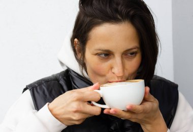 Güzel bir esmer kadın kahve içiyor, sabah kapuçinosu, latte, elinde beyaz bir kupa tutan mocaccino. Soğuk havalarda kafede çekici bir kız. Yaşam tarzı rutini.