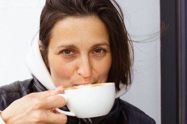 Güzel bir esmer kadın kahve içiyor, sabah kapuçinosu, latte, elinde beyaz bir kupa tutan mocaccino. Soğuk havalarda kafede çekici bir kız. Yaşam tarzı rutini.