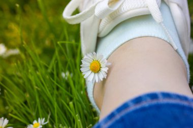 People resting in nature in spring park. A girl in white sneakers sits on green grass on blossoming lawn, a small white daisy flower adorns her leg. Relaxing outdoors in city park, woods. Hello summer