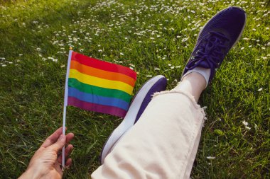 LGBT bayrağı gökkuşağı renginde ve kadın el manzaralı. Lezbiyen, biseksüel, eşcinsel bir kız güneşli bir günde yeşil açıklıkta rahatça oturur. Haziranda Gurur Ayı. Piknikteki yüzü olmayan kadın. Kot pantolonlu bacaklar.