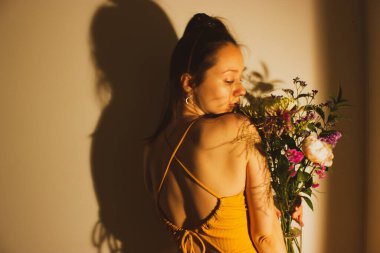 Sırtında açık elbisesi olan ve çiçek buketi kokan güzel kız. Koyu gölgeli seksi kadın. Yazlık elbiseli esrarengiz bayan. Kadın güzellik konsepti. Kutlama, tatil için tebrikler..