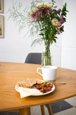 Konforlu mutfak kompozisyonu. Bir tabak domates, dilimlenmiş ekmek, beyaz seramik bardak, beyaz bir duvara karşı çiçek buketinin yanındaki ahşap oval masada duran bardak. Evde yemek yemek. Lezzetli sağlıklı yemek..