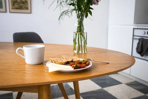 Sıcak ev mutfağı. Bir tabak vişneli domates, dilimlenmiş ekmek ve beyaz bir seramik kupa beyaz bir duvara karşı duran bir buket çiçeğin yanındaki ahşap oval masada duruyor. Evde yemek yemek. Lezzetli sağlıklı yemek.