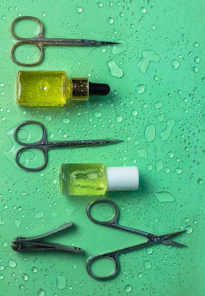 Manicure pedicure items set on vertical green background with water drops. Manicure scissors, tweezers, cutters, cuticle nail growth oil. Beauty shop goods. Nail care cosmetic tools. Spa treatments.