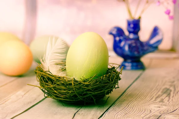 Green painted Easter egg in birds nest with white feather on wooden table. Rustic Easter background. April holidays postcard. Easter eggs wallpaper. Vase in the form of a blue bird on a background.