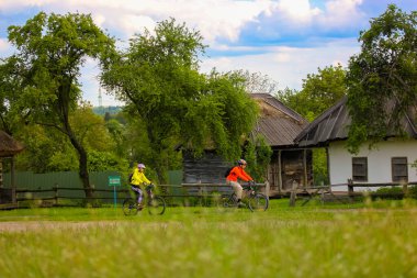 Kyiv, Ukrayna. 1 Mayıs 2021 İki bisikletçi yeşil bir bahar çayırında bisiklet sürüyor. Arka planda geleneksel Ukrayna eski evleri. Ukrayna konseptine yolculuk. Dünyayı dolaş. Yaz sporu