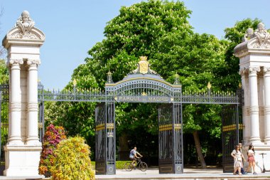 Madrid, Spain. 7 May 2022. Black metal gate with white columns at entrance to Retiro Park. Botanical Garden on a sunny spring day when the trees are in bloom. People walking outdoors, riding bicycles.