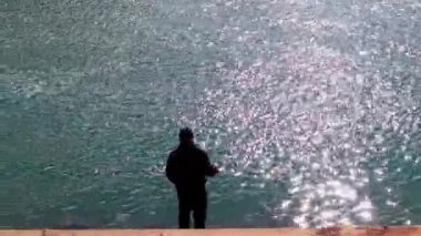 Bir balıkçının silueti elinde olta ile bir gölün ya da nehrin kıyısında hızlı akan bir suyla balık tutuyor. Sportif hobi. Avlanma ve balık tutma. Bahar güneşli bir günde balık tutan adam. Suyun üzerinde güneşin parlaklığı.