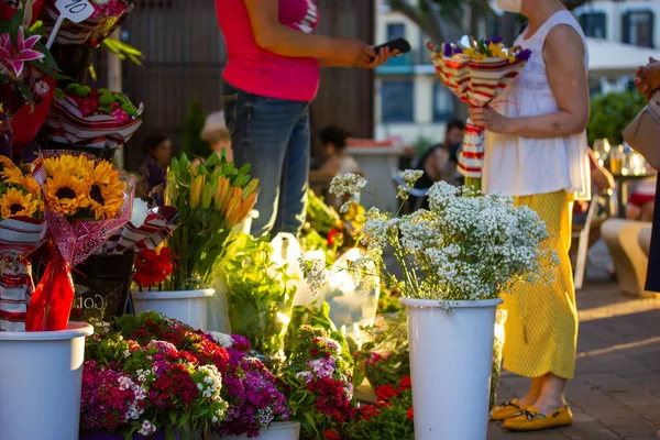 Flower store on a street. Woman buying bouquet of flowers, paying cashless to florist vendor. Many different seasonal flowers in plastic vases stand on a sidewalk in summer day. Urban life scene.