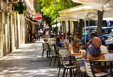Madrid, İspanya. 1 Haziran 2022. İspanyol caddesi, teraslı kafe, restoran. Birçok insan güneşli bir yaz gününde ağaçların gölgesinde bir şehir caddesinde masalarda oturarak rahatlıyor. Şehirde hafta sonu..