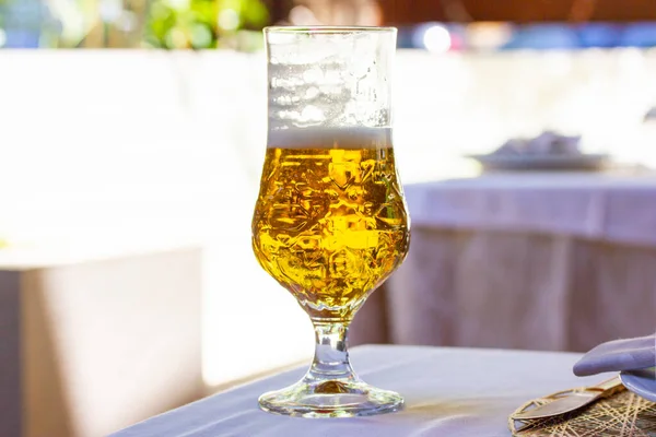 A beer in Tulip glass. Beautiful crystal glass with golden freshly brewed cold wheat beer stands on white tablecloth on table on cafe, restaurant terrace. A refreshing alcoholic beverage in summer day