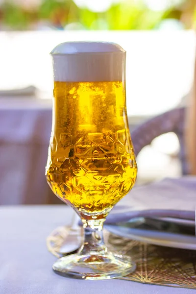 Amber beer in tulip glass. Crystal elegant tableware with golden freshly brewed wheat beer stands on white tablecloth on table in cafe, restaurant terrace. Refreshing alcoholic beverage in summer day.