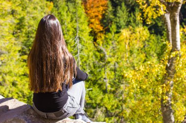 Dağın tepesinde uzun siyah saçlı bir kızın yeşil kozalaklı bir ormana karşı oturması. Kimliği belirsiz genç bir kadın ormanda tek başına yürüyüş yapıyor. Macera, gezi konsepti.