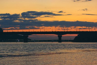 Güneşin batışında, alacakaranlıkta, akşam karanlığında güzel bir kontrastlı köprü silüeti. Rengarenk sarı-kırmızı-mavi bulutlu gökyüzü. Ukrayna, Kiev Panoraması. Dinyeper nehrinde sakin su dalgaları