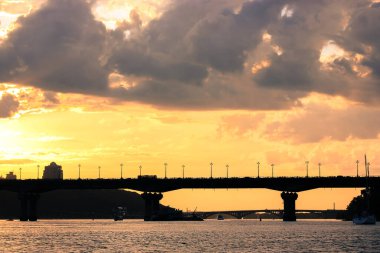 Akşam alacakaranlıkta gün batımında araba köprüsü silueti. Sarı turuncu bulutlu gökyüzü. Ukrayna 'nın başkenti Kyiv' in Panoraması. Dnipro nehrinde sakin bir su. Güzel deniz manzarası. Şehir manzarası.