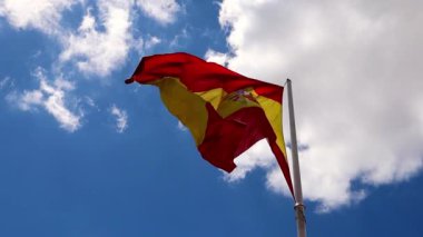 A big red yellow bright Spanish flag with emblem flutters isolated in a wind against a blue sky with white clouds on a sunny summer day. National symbol, patriotism. Slow motion video. Spain, Madrid. 