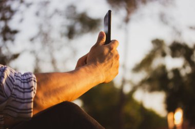 Cep telefonu güçlü adamların elinde. Kimliği belirsiz erkek, akıllı telefon aleti kullanan adam güneşli bir günde parkta oturuyor. Sokakta telefon kullanmak. İnternet bağlantısı. Kablosuz internet açık havada. Cep telefonuyla fotoğraf çekiyor.