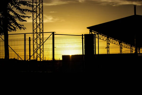 Contrast silhouette of an industrial building behind lattice fence against the evening sunset yellow sky. Warehouse, factory building roof. Commercial property exterior. Wire fence in industrial zone.
