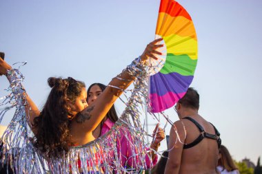 Madrid, Spain. July 25, 2022 People have fun at a festival at Pride Month. Gay Parade 2022 in summer. Merry people having fun dancing. Young woman with rainbow LGBT fan. Glitter, tinsel party outdoors