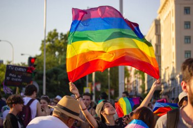 Madrid, Spain. July 25, 2022 Cheerful teenage girl, young woman holds a rainbow LGBT flag above her head at a gay parade, festival at Pride month. Fun, joy, proud, youth freedom entertainment concept.