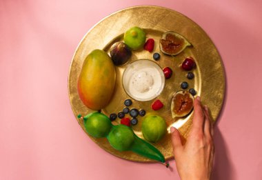 Woman's hands reaches for served plate, platter of fresh tropical fruit on a pink table top view. Faceless girl serving on a table. Figs, mango, berries, cherries, limes, ice cream, yogurt on a tray.