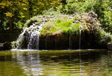 Artificial stone fountain on a small pond. Water flows in a lake, mountain river. A natural landscape in a green public state park, forest, square against green trees. Calm aqua in a water reservoir. 