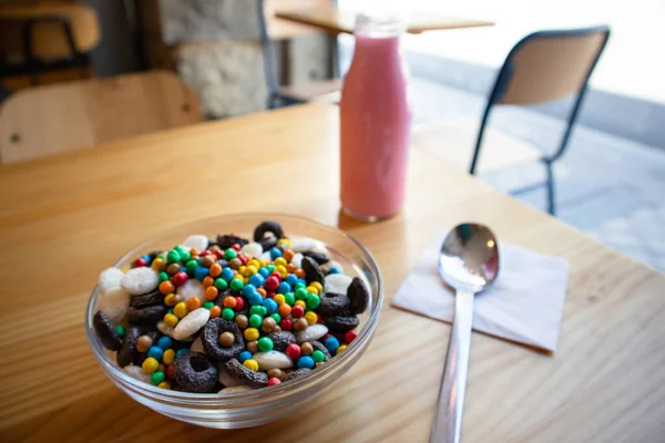 Dry breakfast in glass round plate bowl on a wooden table in cafe. Delicious cornflakes and pink milk in a bottle. Useful healthy breakfast. Food for kids. Different type cereals, multicolored balls.