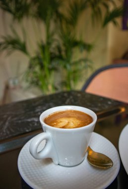 Porcelain white coffee cup with cappuccino, mochaccino on a saucer with a teaspoon on a black marble table to view. Hot tasty drink for a breakfast, branch, lunch. Aromatic beverage on a kitchen.